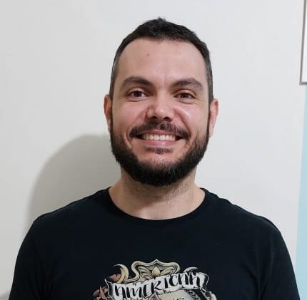 Bruno é homem branco, de cabelos curtos castanhos e barba. Veste camiseta e calça pretas.